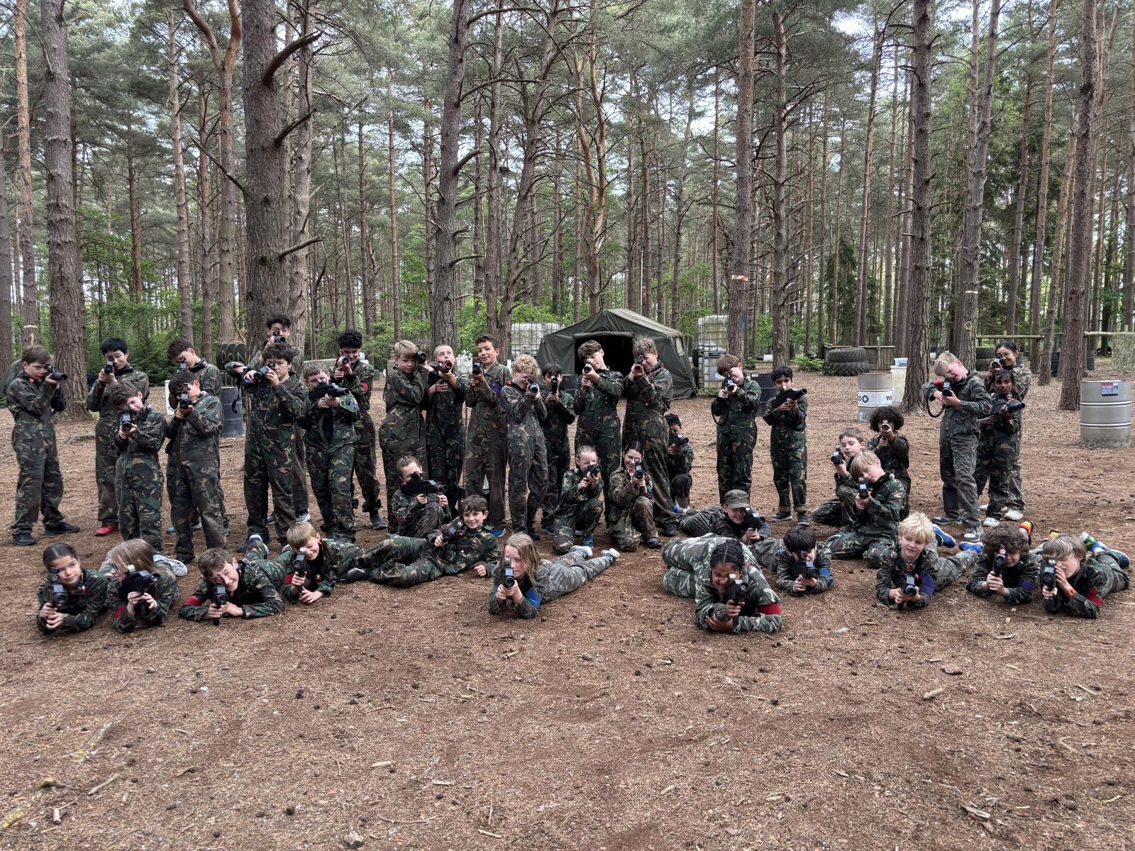 A large group of children in camouflage overalls aiming laser rifles in pine woodland